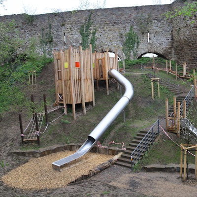 Das Bild zeigt einen Spielplatz an einem Steilhang mit zwei Rutschbahnen, einem Holzturm, einer Treppe und Sitzsteinen.