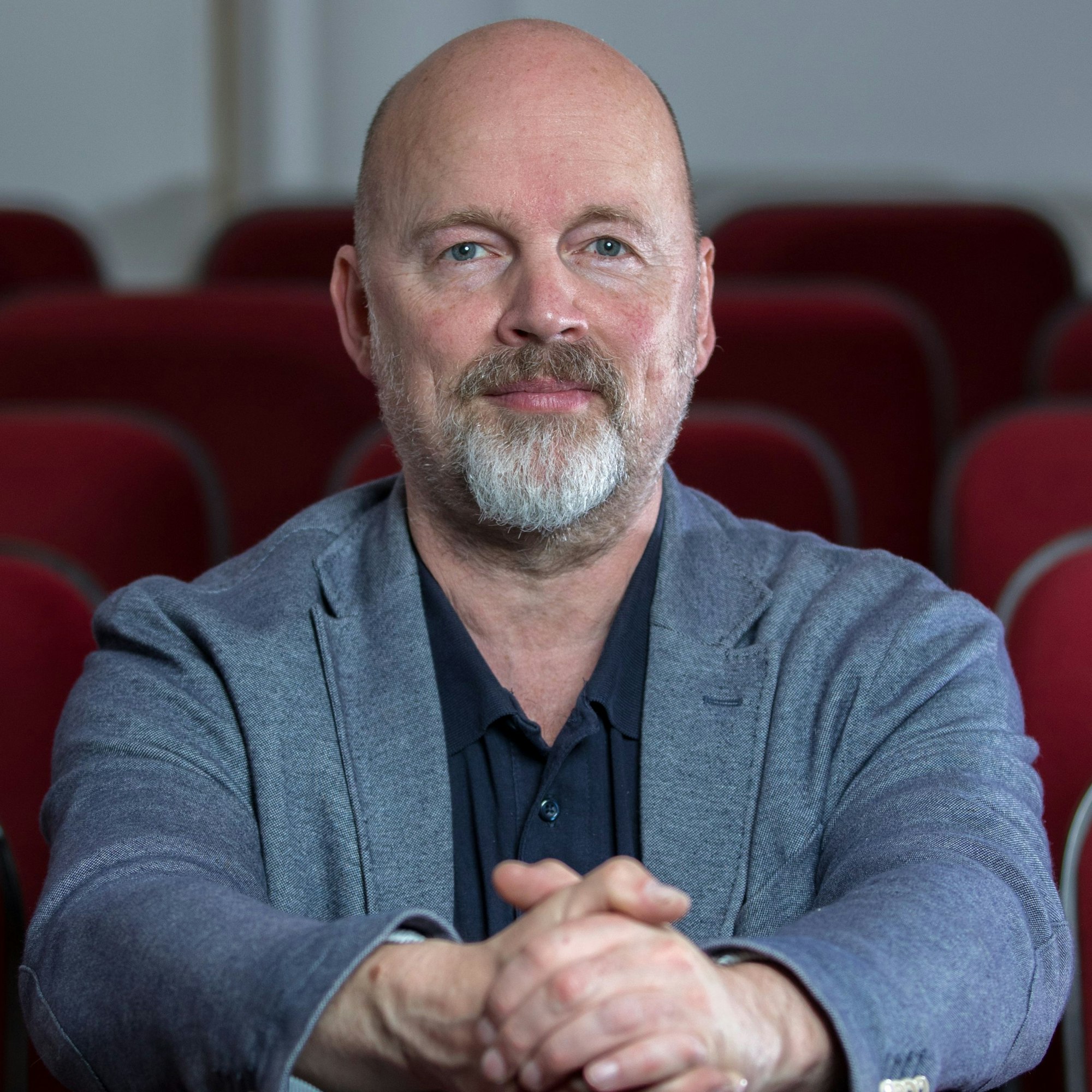 Ein Mann mit blauem Sakko, im Hintergrund rote Sitzplätze im Theater.