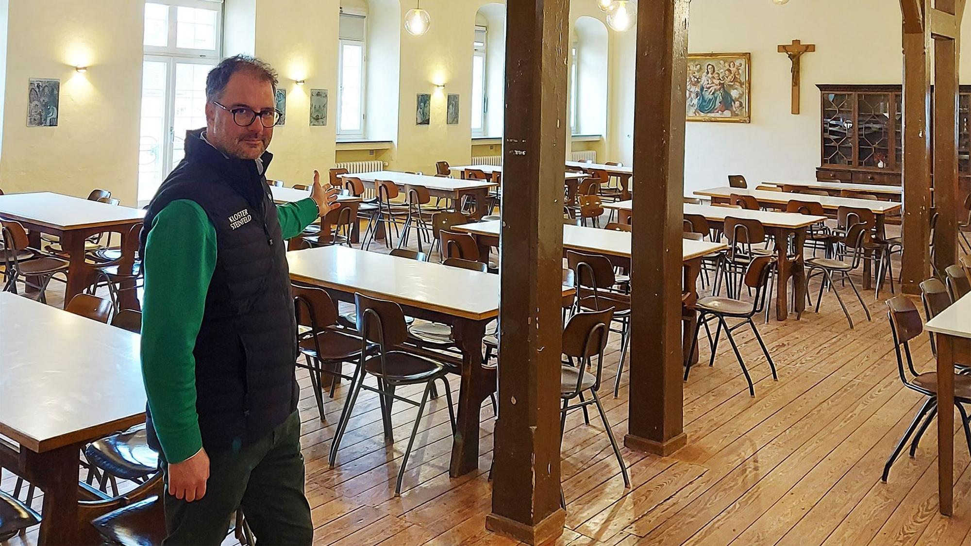 Christoph Böhnke zeigt in den Hermann-Josef-Saal im Kloster Steinfeld, in dem zahlreiche Tische und Stühle stehen.