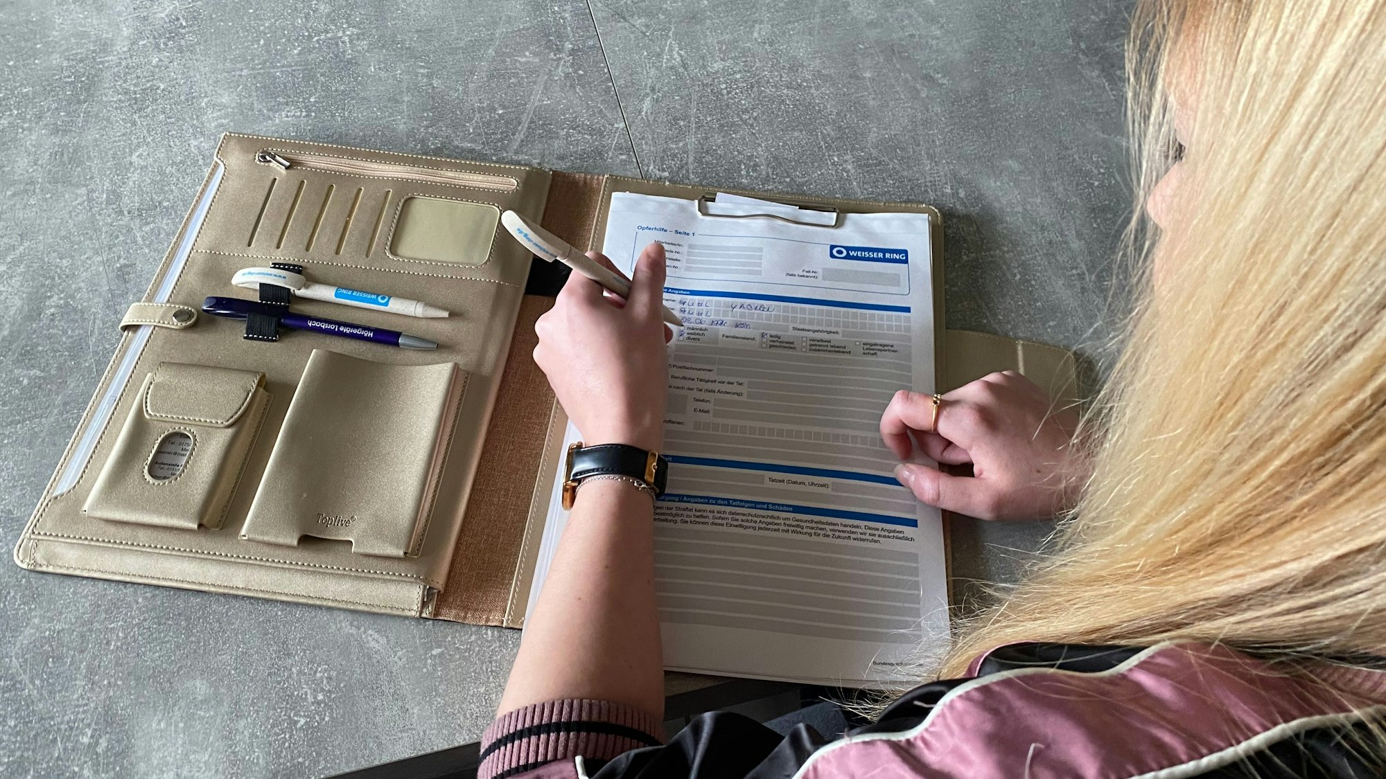 Auf einem Tisch liegt eine beigefarbene Mappe. Darin befinden sich Formulare des Weißen Rings. Yasmin Fuhl füllt die Formulare mit einem Kugelschreiber aus.