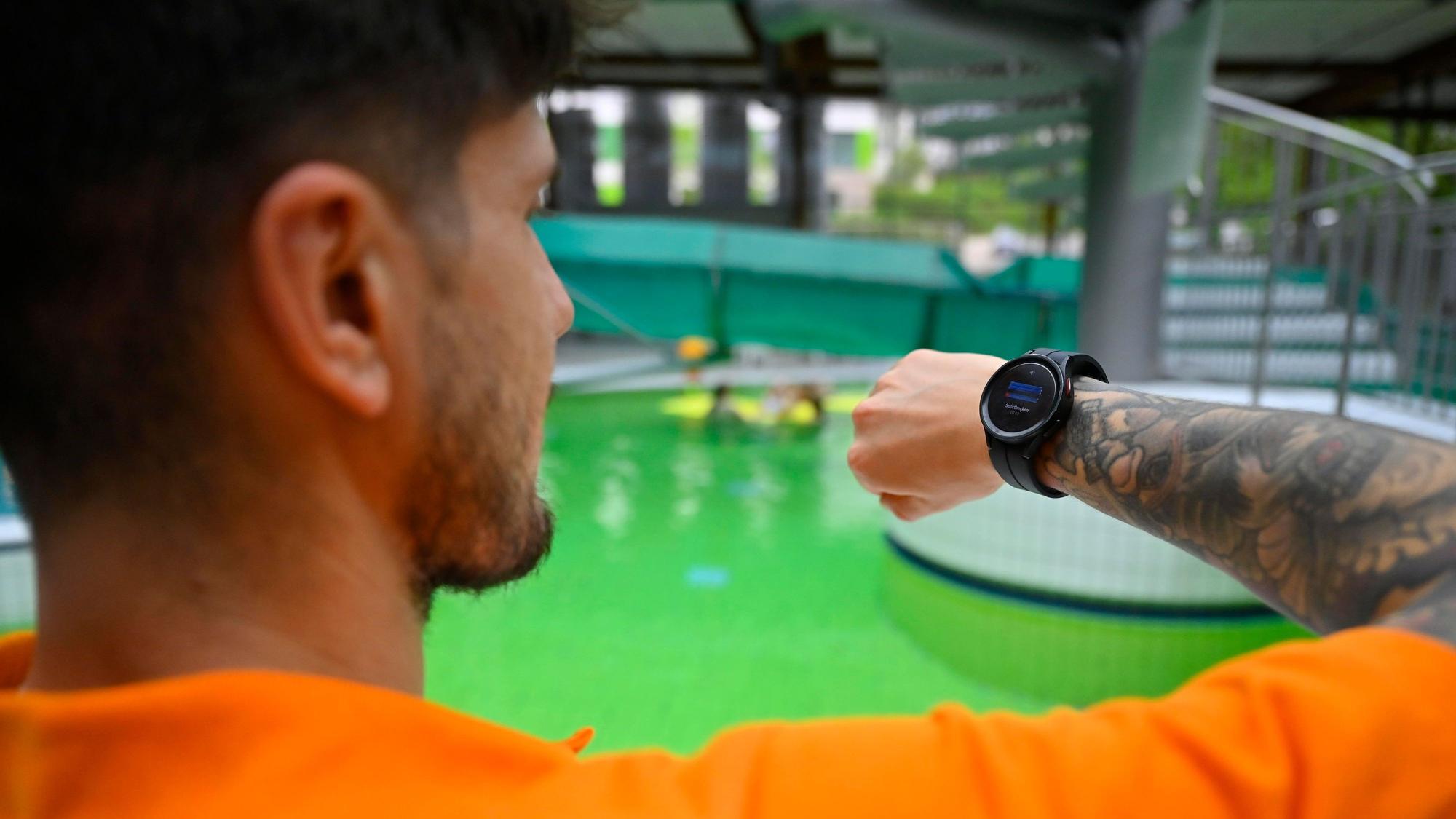 Ein Mann steht am Rand eines Schwimmbeckens und schaut auf eine Smartwatch auf seinem Handgelenk.