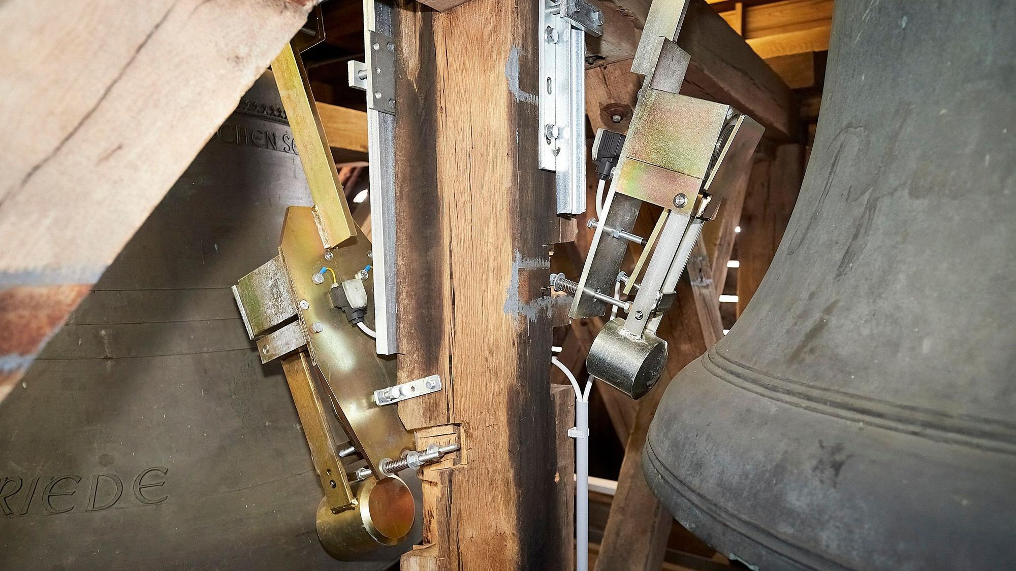 An einem alten Glockenstuhl aus Holz sind neue, pneumatische Schlagwerke angebracht, die eine Glocke, die zum Teil zum rechts im Bild zu sehen ist, zum Klingen bringen.