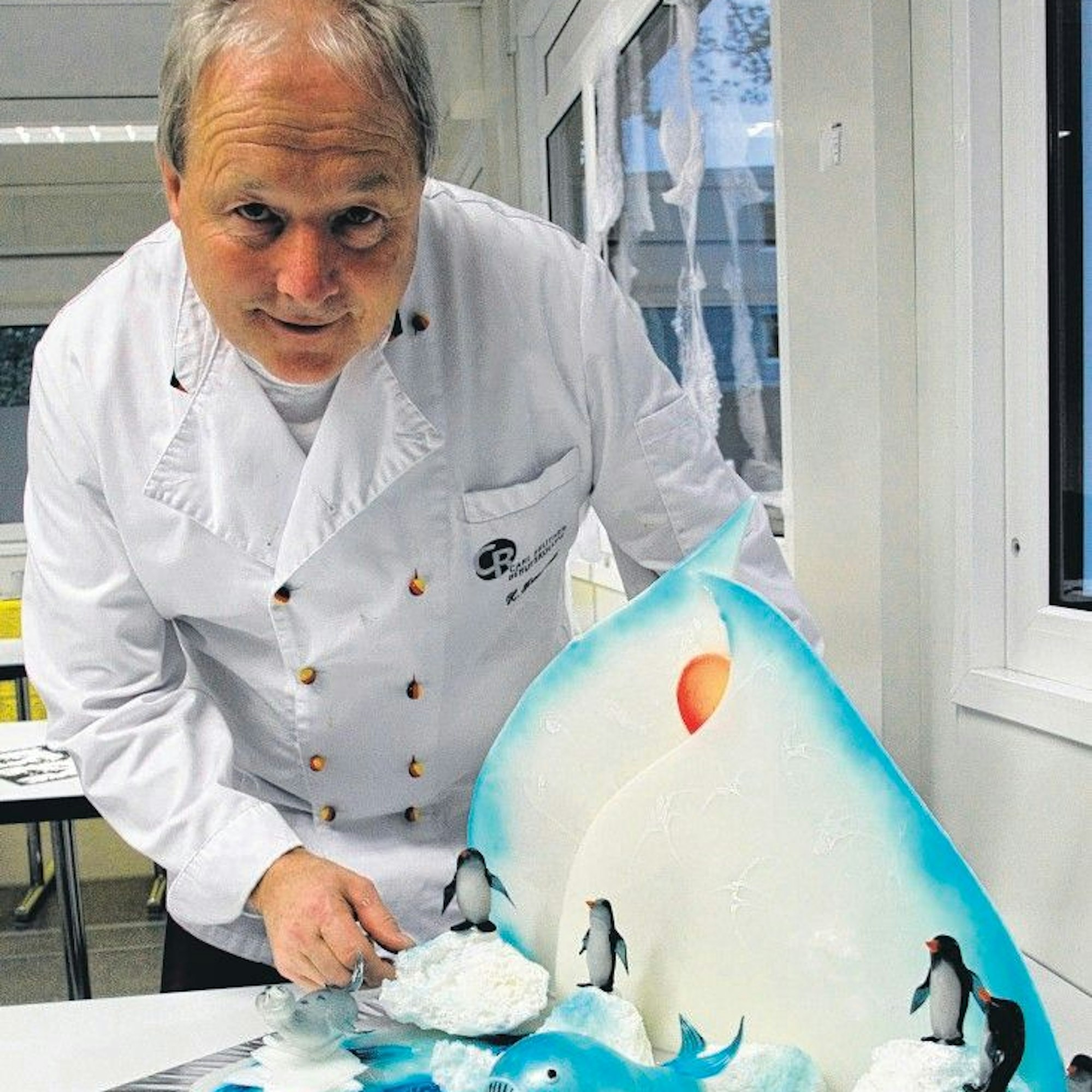 Das Bild zeigt Heiner Krautscheid in weißer Konditorjacke. Vor ihm steht eine aus Zucker geformte Südpol-Szenerie mit Eisberg, Eisschollen, Pinguinen und einem Wal.