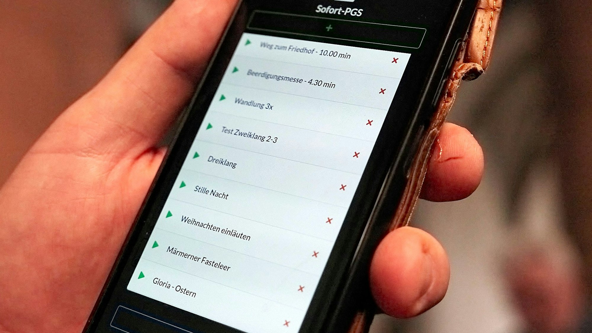 In einer Hand wird ein Smartphone gehalten, auf dessen Bildschirm verschiedene Optionen zu sehen sind. Sie können gewählt werden, um die Glocken der Kirche in Marmagen zu steuern.