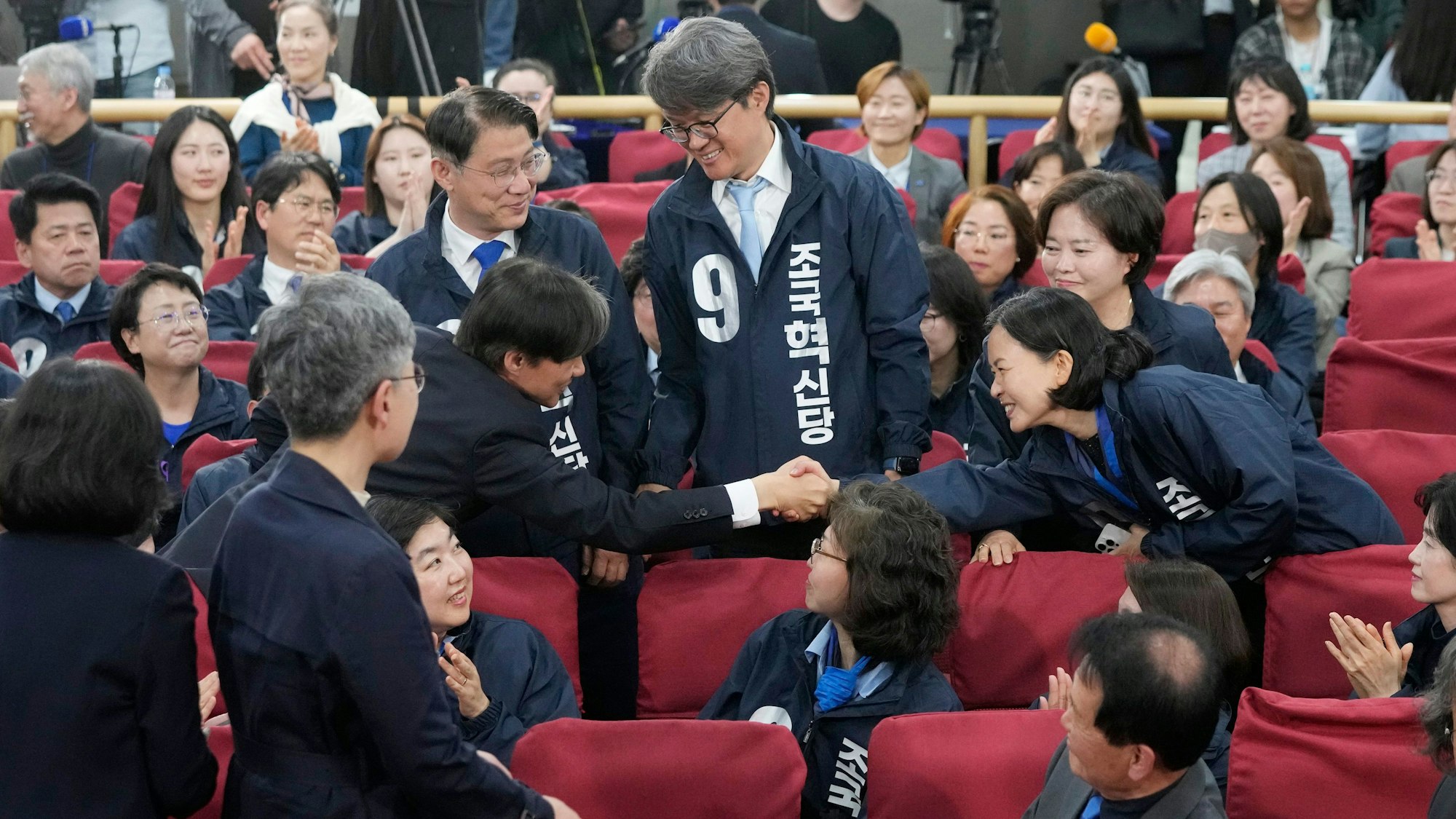 Mehrere Personen sitzen auf Stühlen und sind in Richtung eines Mannes in der Bildmitte gewandt, dem die Hand geschüttelt wird.