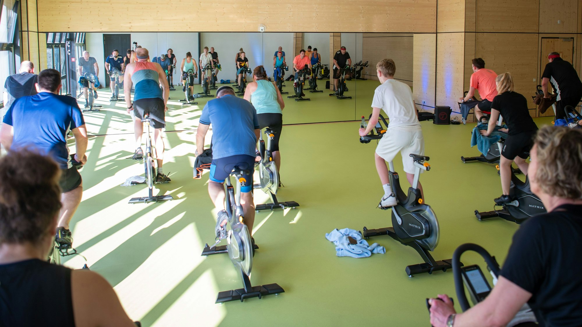 Mehrere Menschen sitzen auf Cycling-Bikes in einem großen Raum vor einem Spiegel.