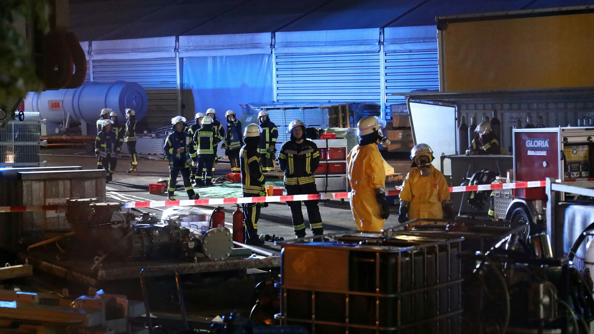 Feuerwehrleute stehen auf dem Werksgelände, ein Teil ist mit einem rot-weißen Flatterband abgesperrt.