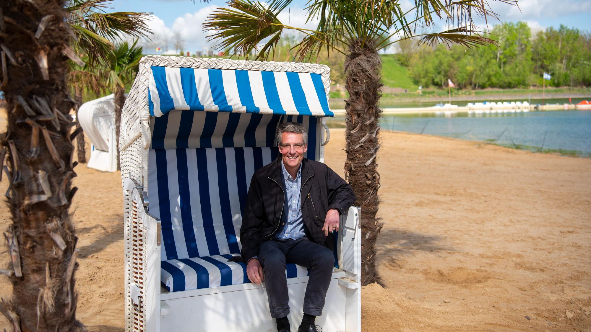 Geschäftsführer Christoph Hartmann sitzt in einem Strandkorb unter Palmen am Badestrand des Zülpicher Sees.