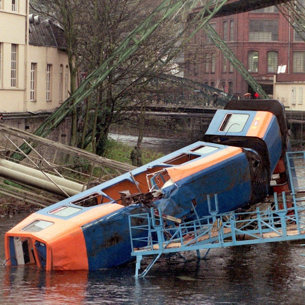 Der abgestürzte Zug der Wuppertaler Schwebebahn im Stadtteil Elberfeld liegt zerborsten in der Wupper. Hoch über der Bahn, in der ein großes Loch klafft, sieht man das Fahrgerüst.