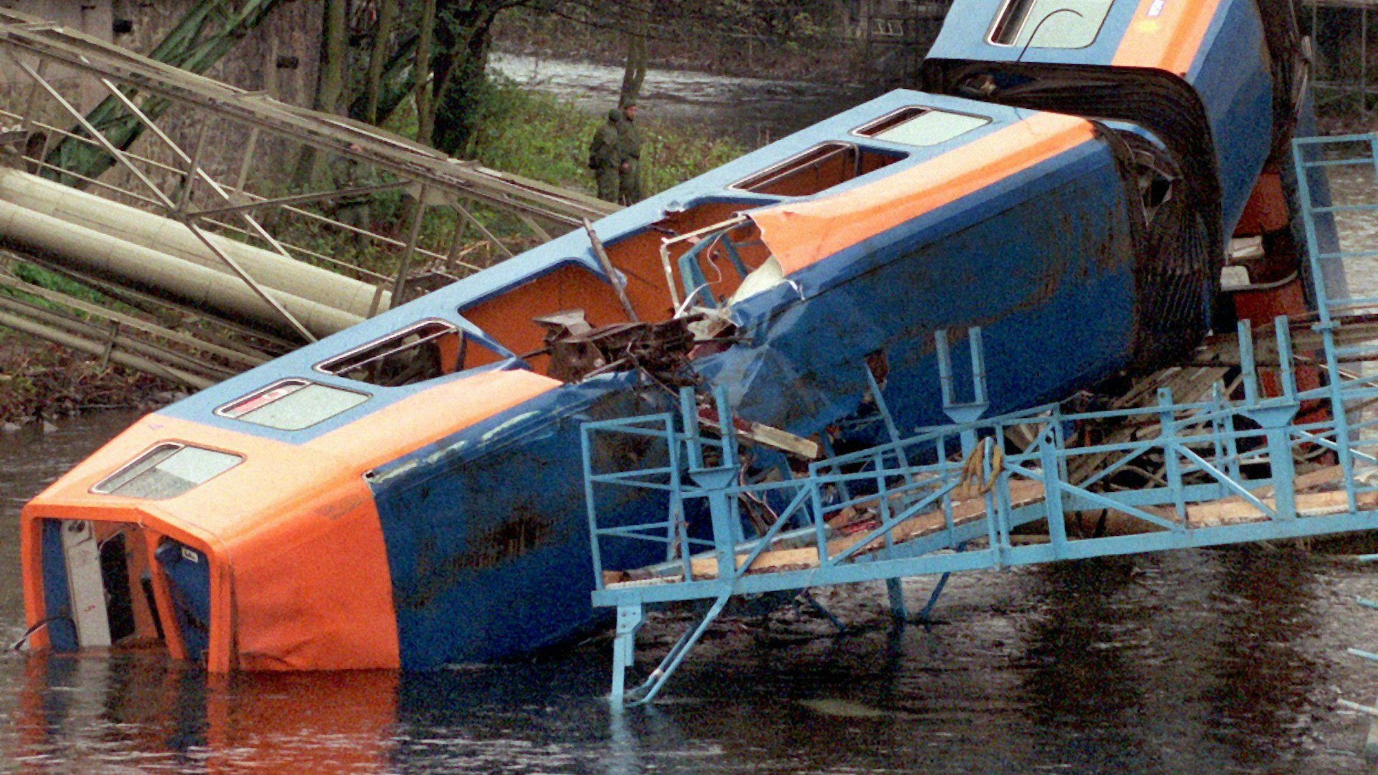Der abgestürzte Zug der Wuppertaler Schwebebahn im Stadtteil Elberfeld liegt zerborsten in der Wupper. Hoch über der Bahn, in der ein großes Loch klafft, sieht man das Fahrgerüst.