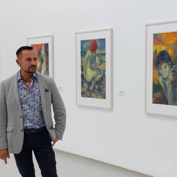 Ein Mann im grauen Sakko und einem weiß-blau gemusterten Hemd in einem Museumssaal; an der Wand hängen farbenfrohe Bilder.