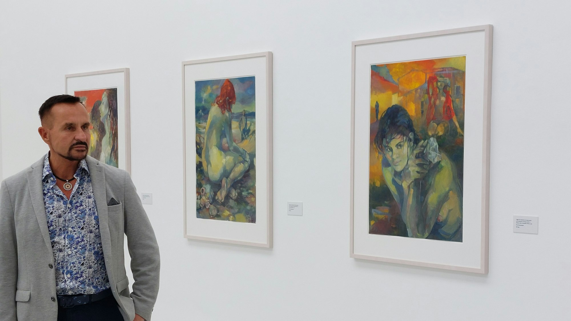 Ein Mann im grauen Sakko und einem weiß-blau gemusterten Hemd in einem Museumssaal; an der Wand hängen farbenfrohe Bilder.