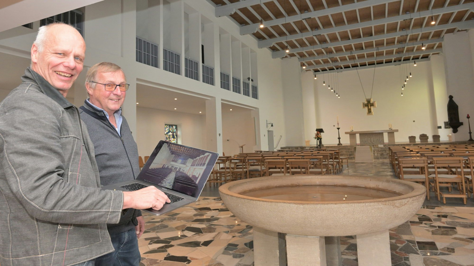 Zwei Männer halten einen Laptop und stehen in einem Kirchenraum mit großem Weihwasserbecken.