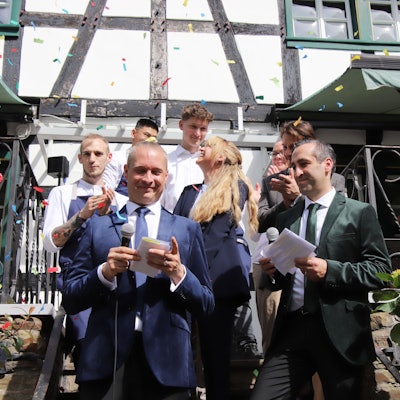 Zwei Männer stehen im Konfettiregen vor einem Fachwerkhaus, Servicepersonal ist hinter ihnen zu sehen.