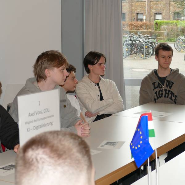 Jugendliche und ein Mann mit grauen Haaren sitzen an einem Tisch, auf dem ein EU-Fähnchen steht.