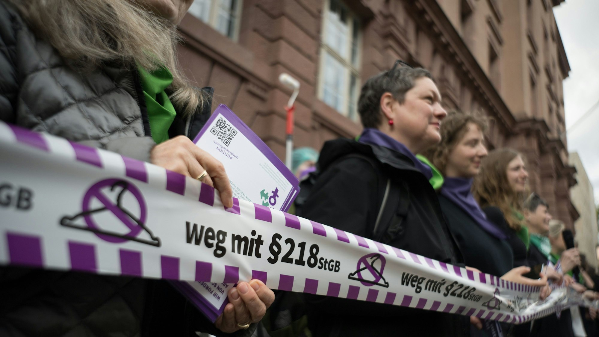 Aktivistinnen halten ein Band mit einem durchgestrichenen Kleiderbügel und dem Schriftzug „weg mit dem §218 StGB“ vor sich.