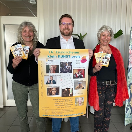 Gaby Bärenklau und Heike Jachtmann halten das Programmheft des 16. Euskirchener Kleinkunstpreises in den Händen. Sie flankieren Sebastian Zimer, der das zugehörige Plakat präsentiert.