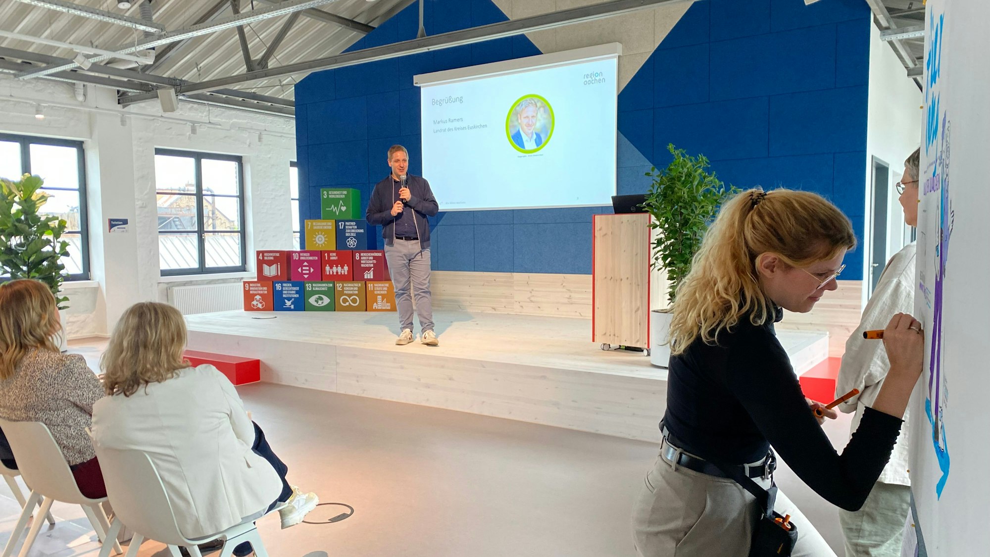 Landrat Markus Ramers steht auf einer Bühne. An einer Tafel malt eine Frau. Im Publikum sitzen zwei Frauen auf weißen Stühlen.