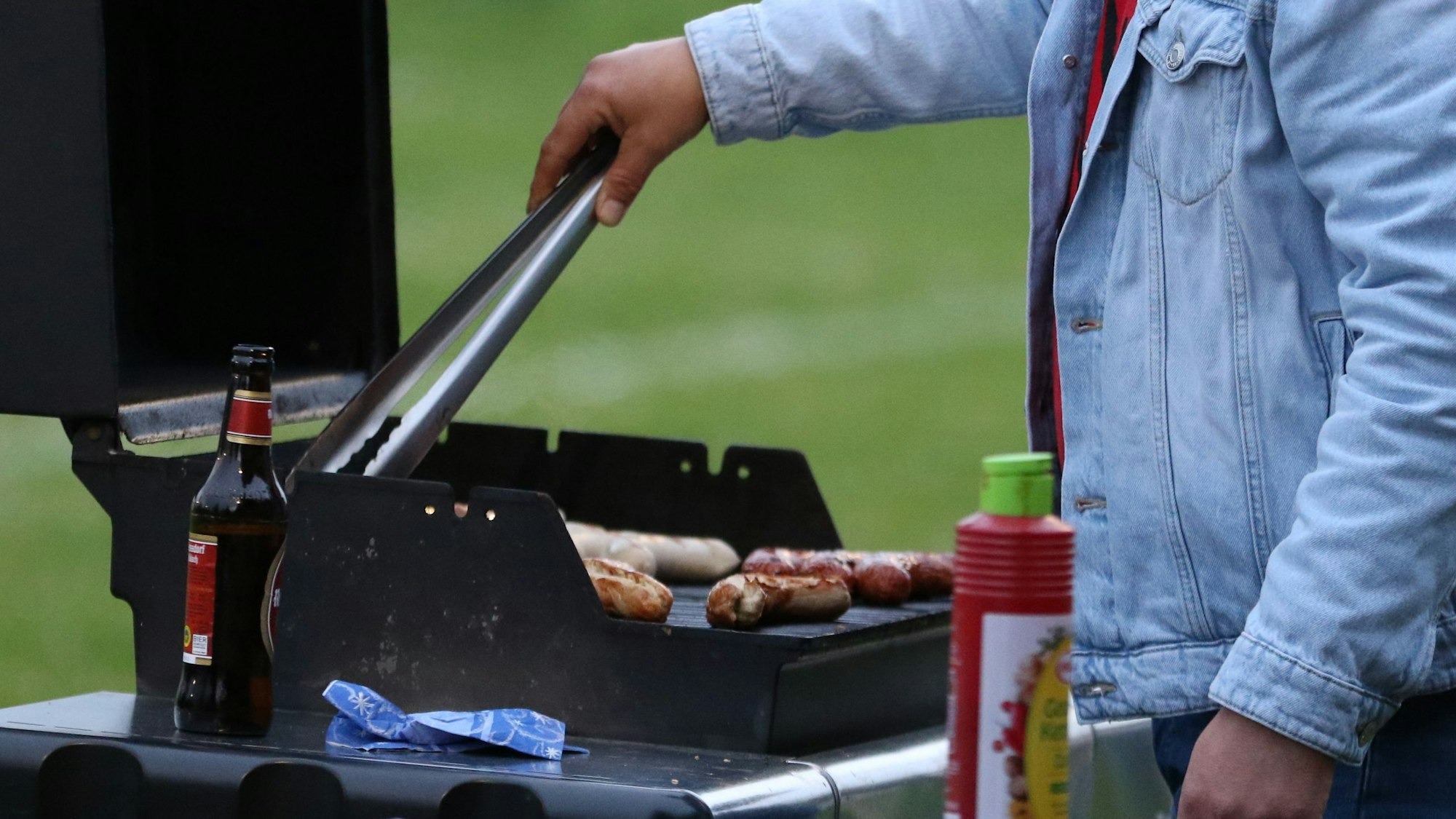 Das Bild zeigt einen Gasgrill. Ein Mann vor dem Grill dreht gerade die Bratwürstchen um.