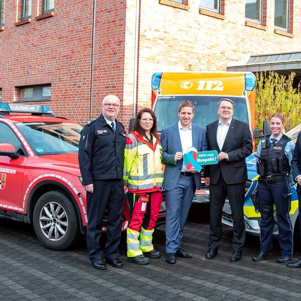 Alexander Rheindorf, Jessica Jeub, Markus Ramers, Udo Becker, Christina Specht und Wolfgang Henseler haben sich zum Gruppenfoto aufgestellt, flankiert von Fahrzeugen der Feuerwehr, des Rettungsdienstes und der Polizei. Ramers und Becker halten ein Respekt-Schild hoch.