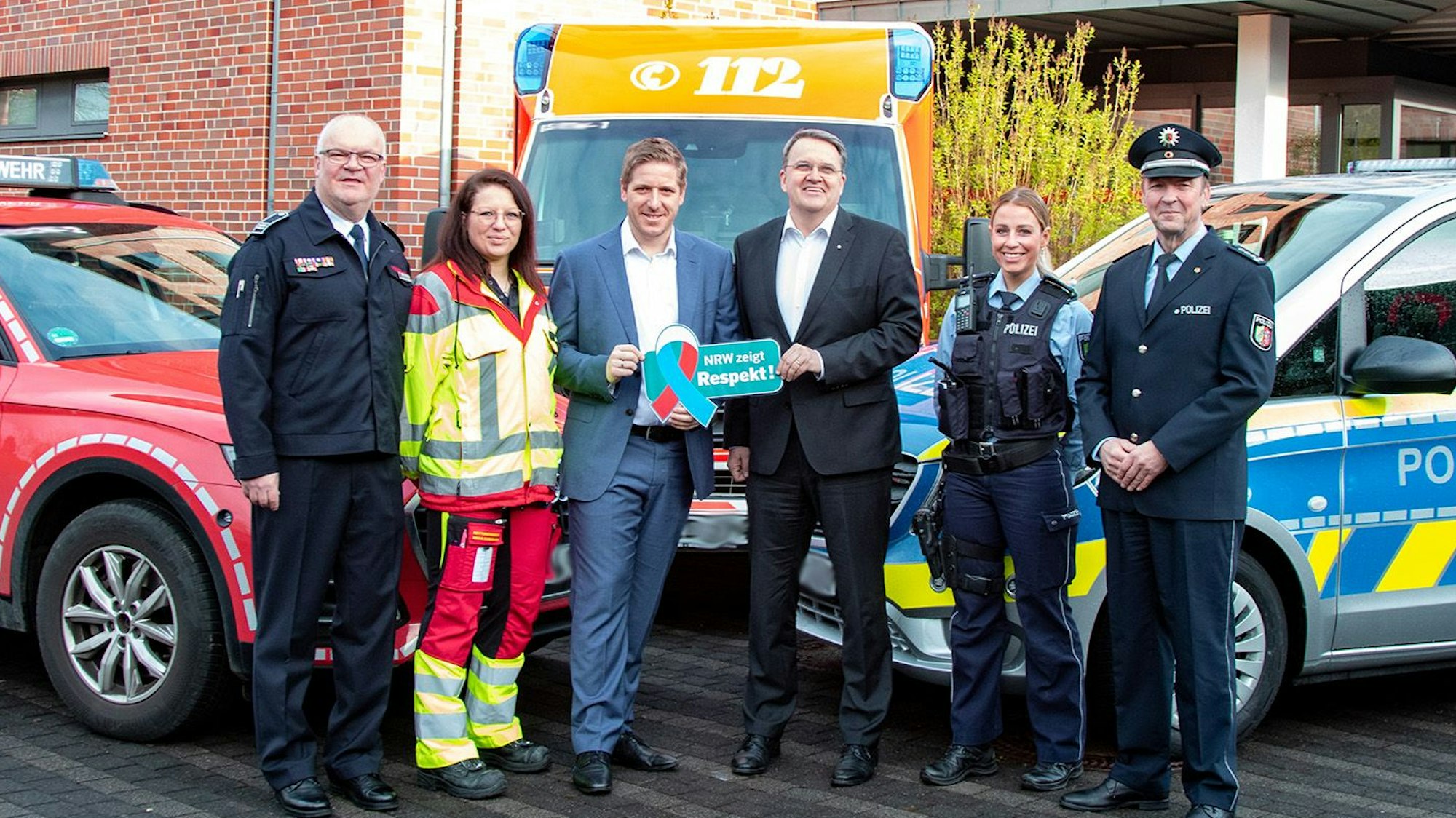Alexander Rheindorf, Jessica Jeub, Markus Ramers, Udo Becker, Christina Specht und Wolfgang Henseler haben sich zum Gruppenfoto aufgestellt, flankiert von Fahrzeugen der Feuerwehr, des Rettungsdienstes und der Polizei. Ramers und Becker halten ein Respekt-Schild hoch.