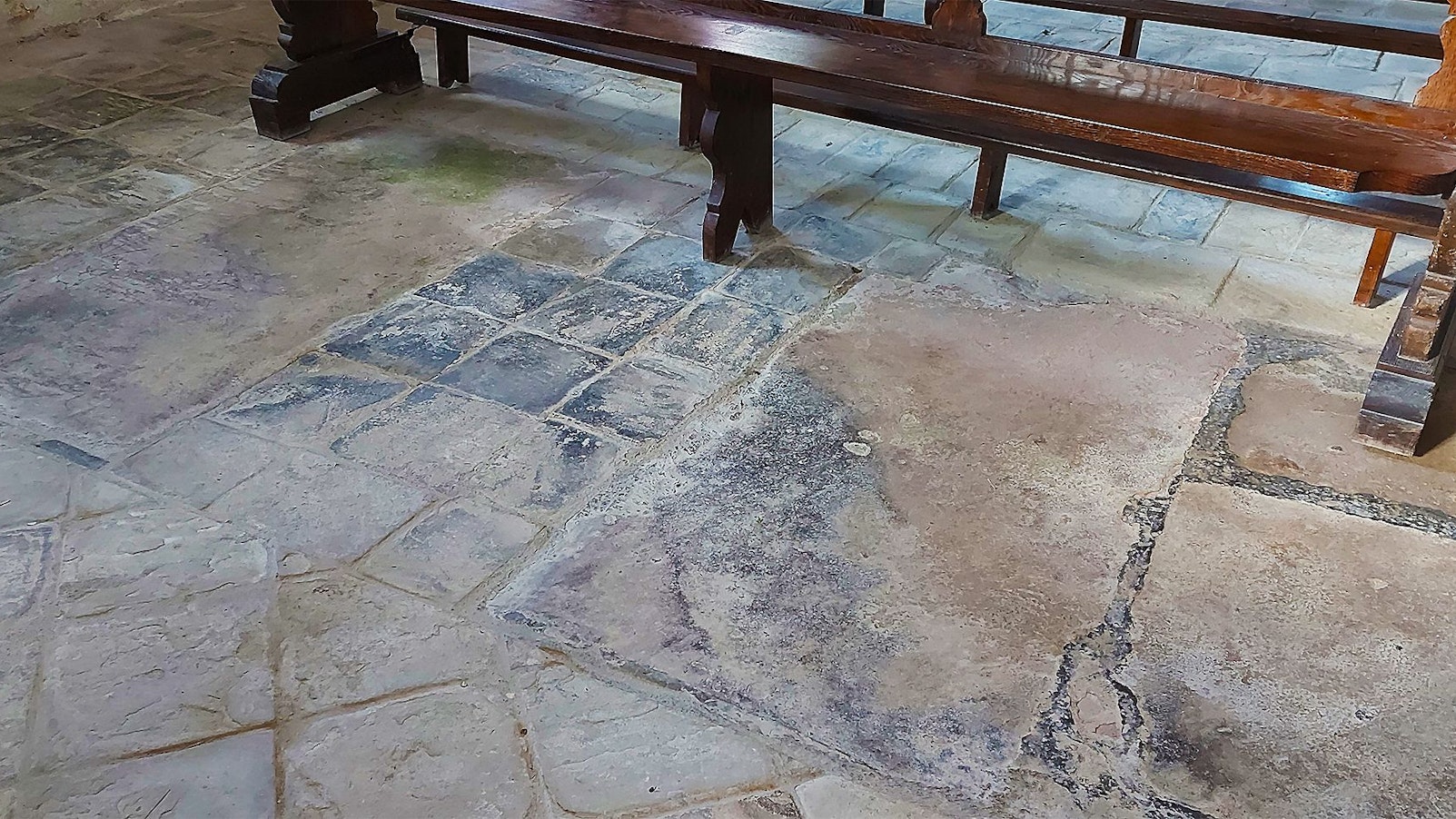 Zwei Grabplatten sind zu sehen, getrennt durch wesentlich kleinere quadratische Sandsteinplatten. Im Hintergrund steht eine Kirchenbank.