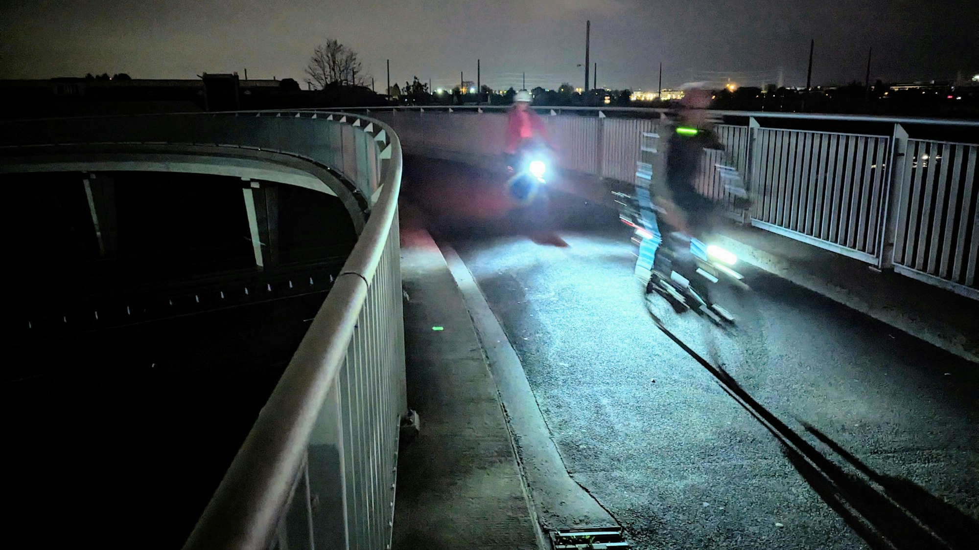 Zwei Radfahrer mit beleuchteten Fahrrädern fahren in der Dunkelheit über eine Brücke.