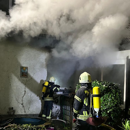 Unter schwerem Atemschutz gehen Feuerwehrleute gegen das Feuer in einem brennenden Haus in Overath-Untereschbach vor.