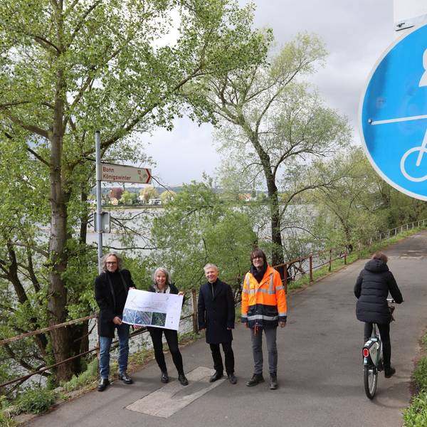Eine vierköpfige Gruppe steht auf einem Radweg, am Rand weisen rot-weiße Schilder nach Bonn und Königswinter..