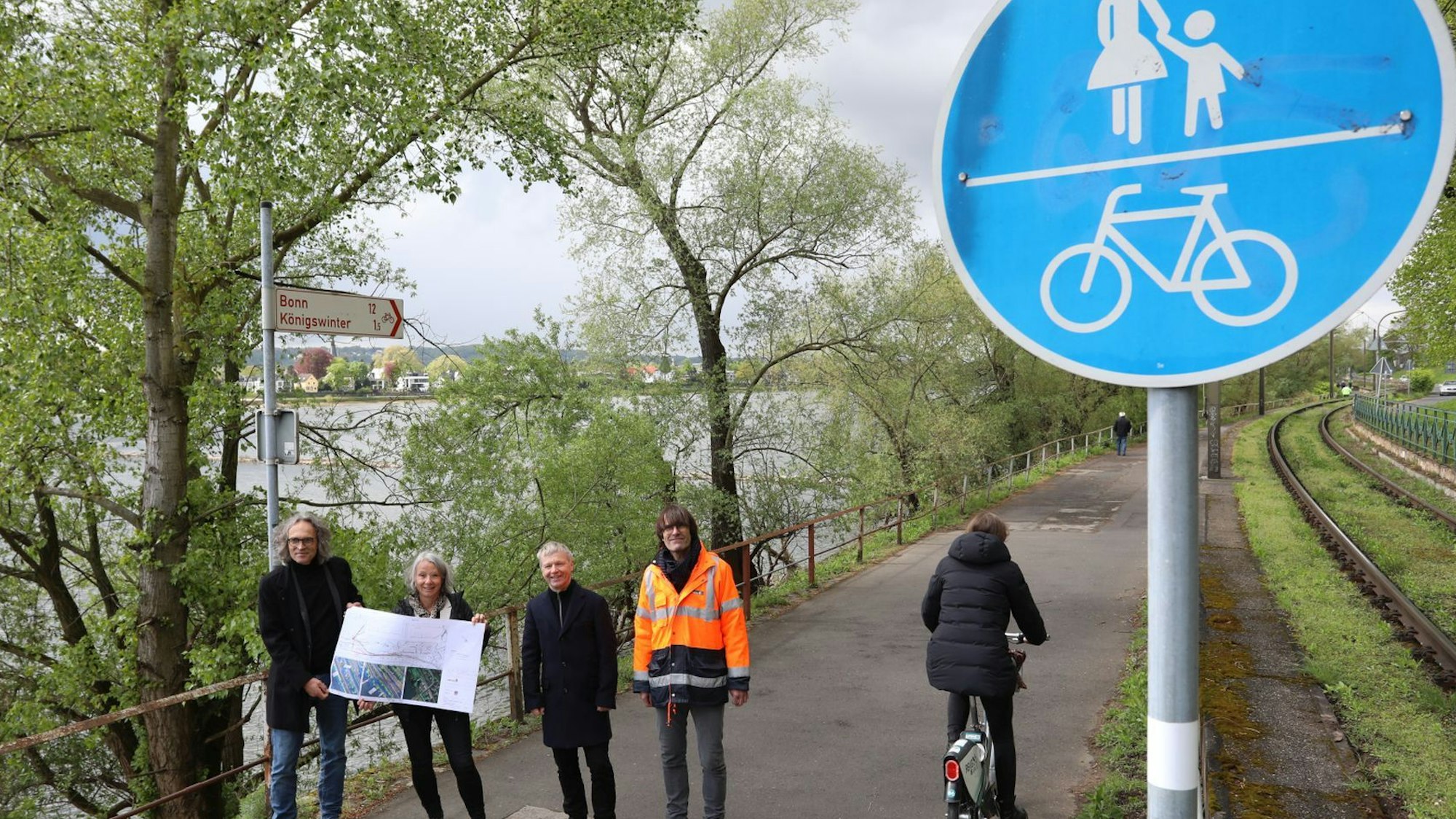 Eine vierköpfige Gruppe steht auf einem Radweg, am Rand weisen rot-weiße Schilder nach Bonn und Königswinter..