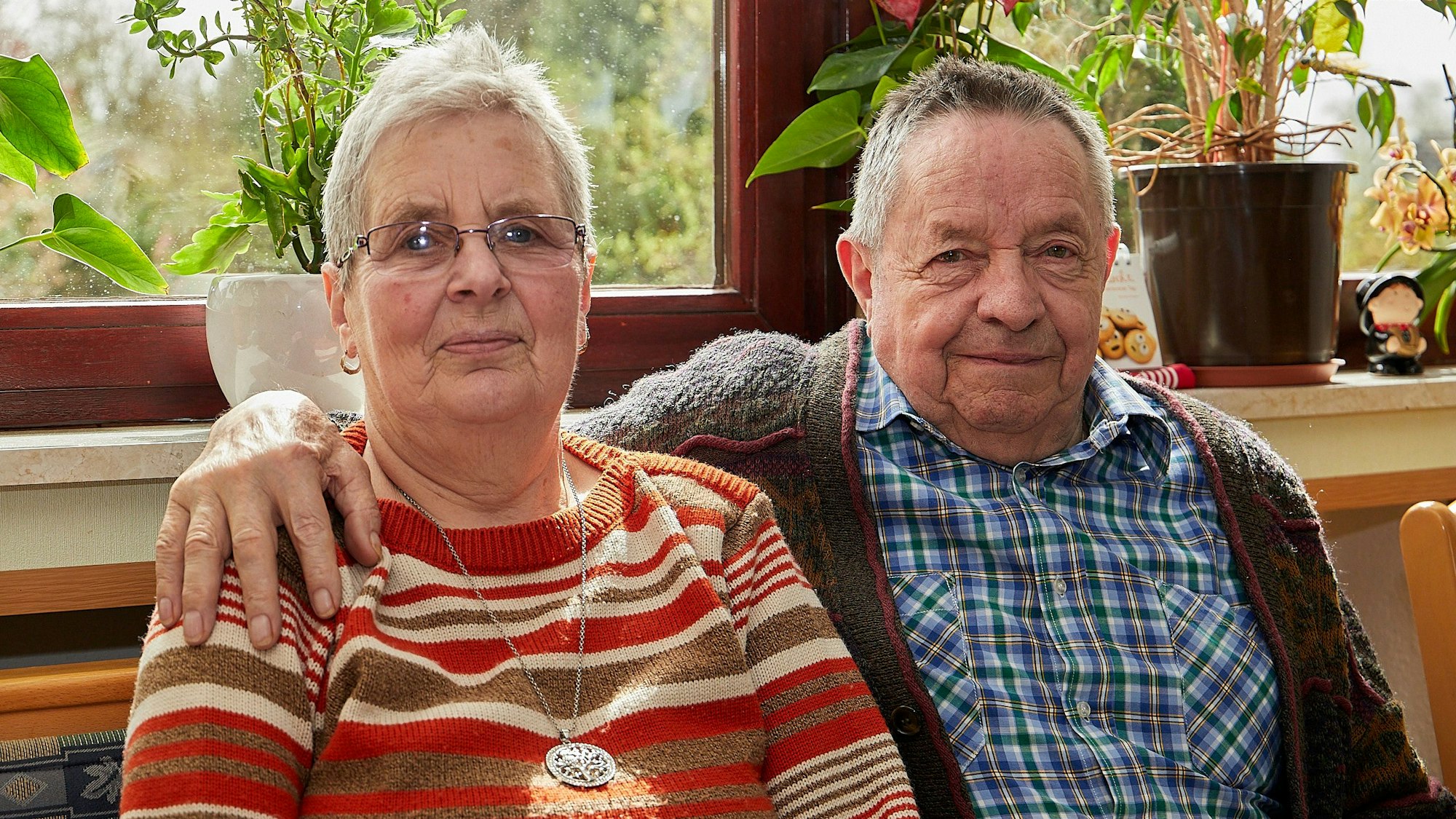 Helene und Klaus Berends sitzen vor einem Fenster, er hat seinen Arm um sie gelegt.