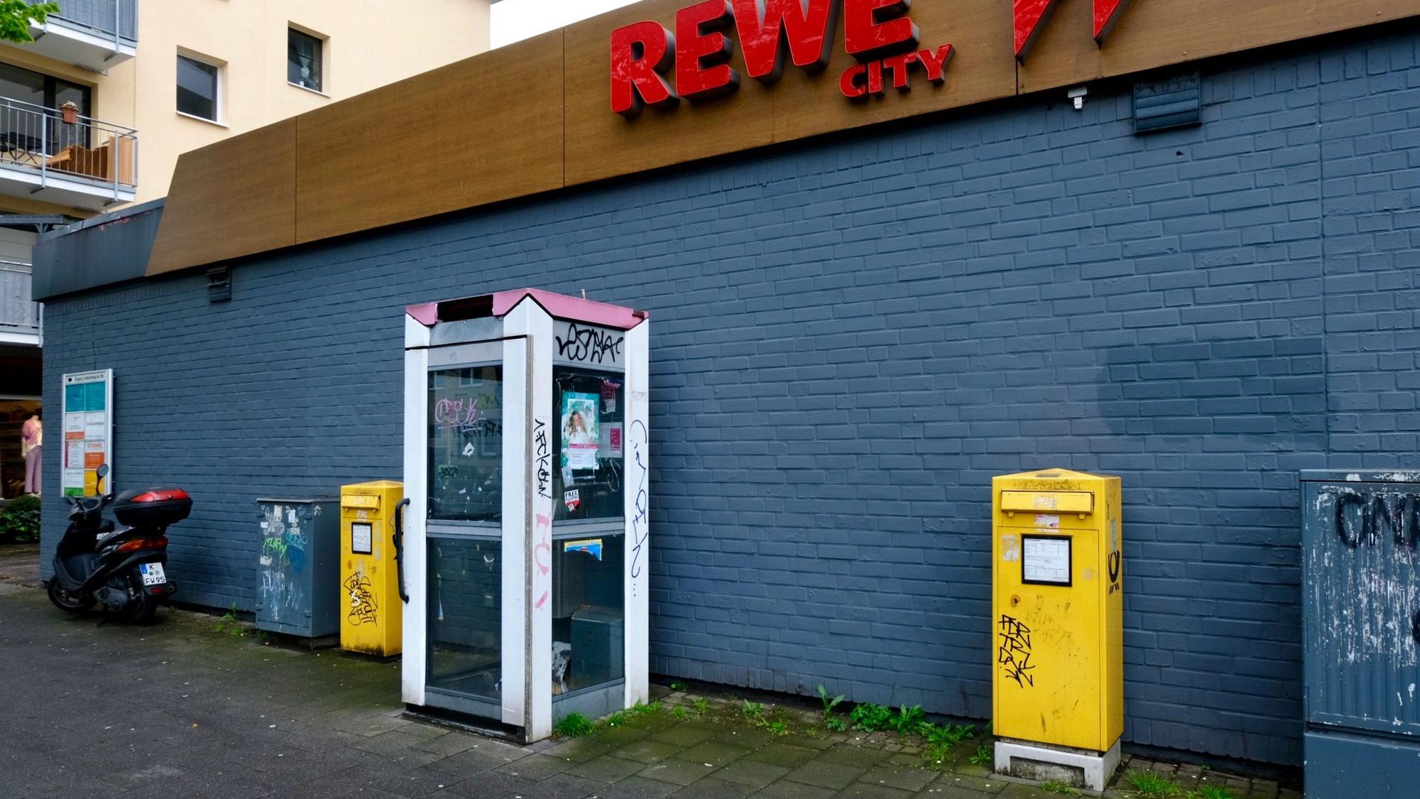 Das Bild zeigt eine verwahrloste und nicht mehr funktionstüchtige Telefonzelle vor dem Rewe-Markt an der Dürener Straße. Foto von Susanne Esch