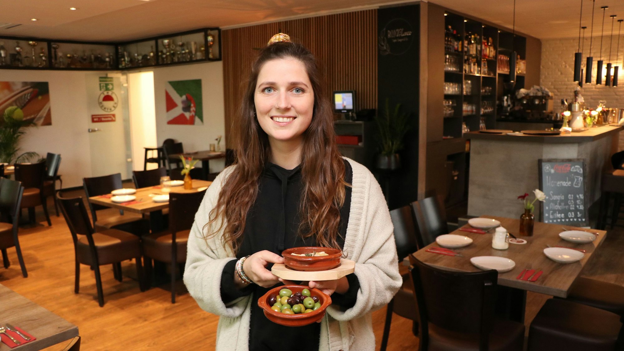 Eine junge Frau steht mit einigen Schalen mit Tapas in den Händen im Gastraum.