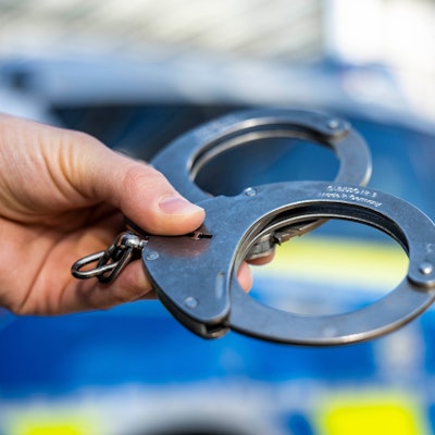 Eine Hand hält Handschellen vor einen Streifenwagen der Polizei.