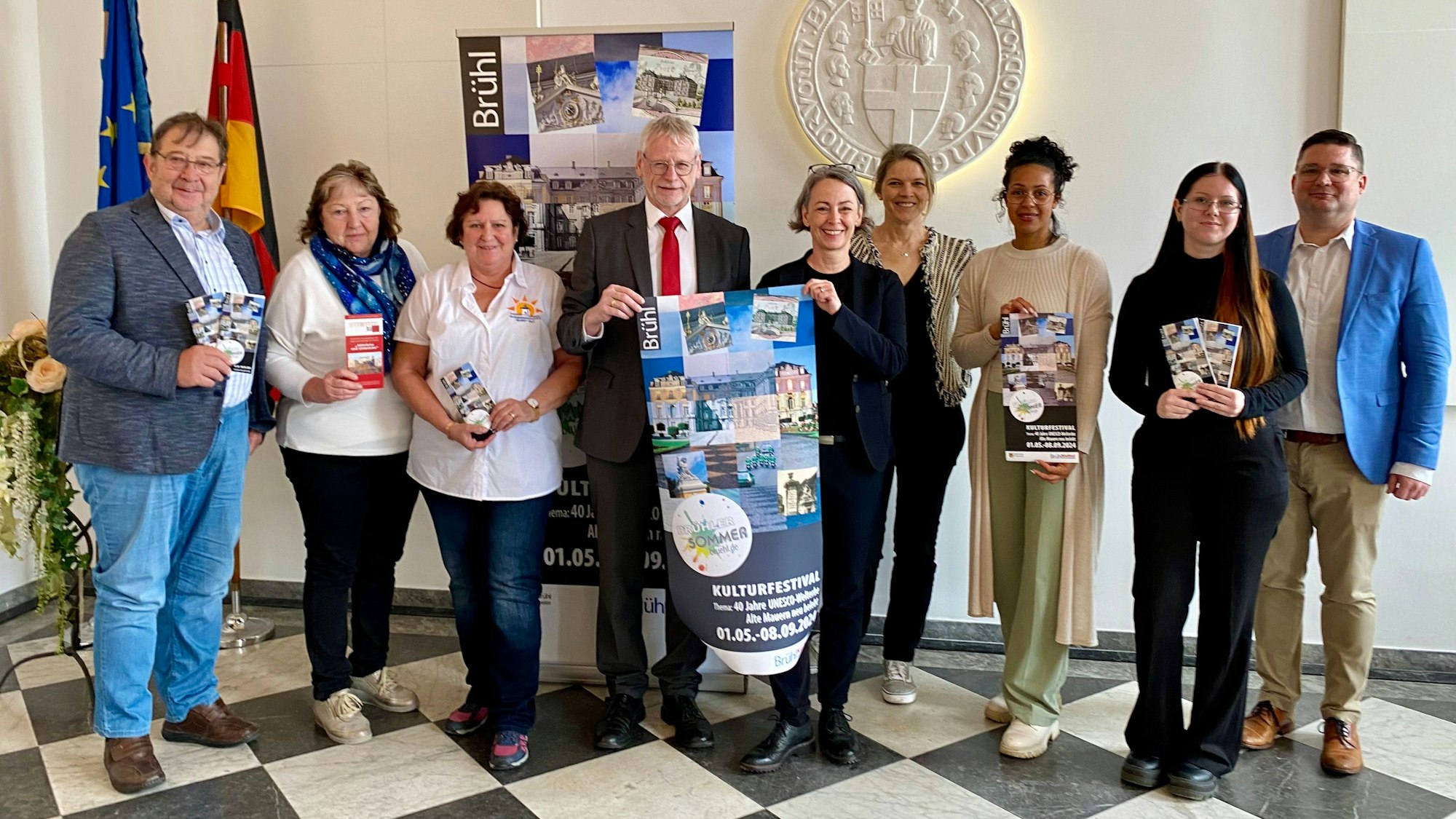 Sechs Frauen und drei Männer lächeln in die Kamera, zwei Personen halten ein großformatiges Plakat mit der Aufschrift „Brühler Sommer“.