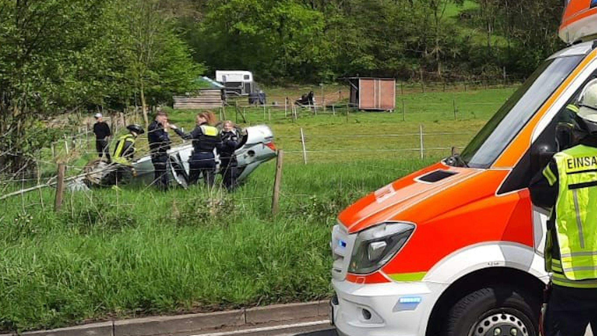 Einsatzkräfte von Polizei und Feuerwehr untersuchen ein Auto, das sich überschlagen hat und auf dem Dach in einer Wiese liegengeblieben ist.