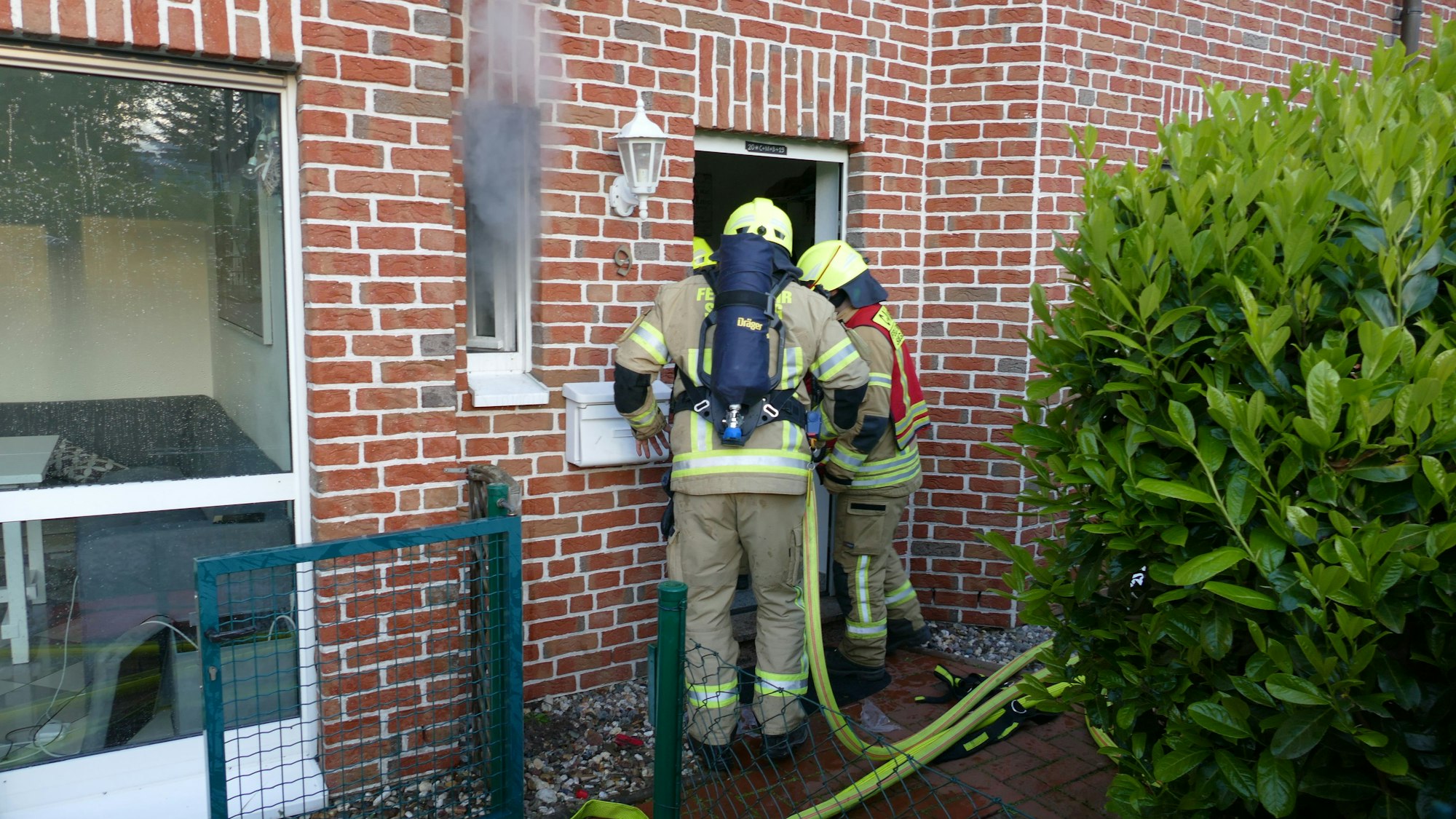 Feuerwehrleute stehen vor einer Haustür, aus dem Fenster daneben kommt Qualm.
