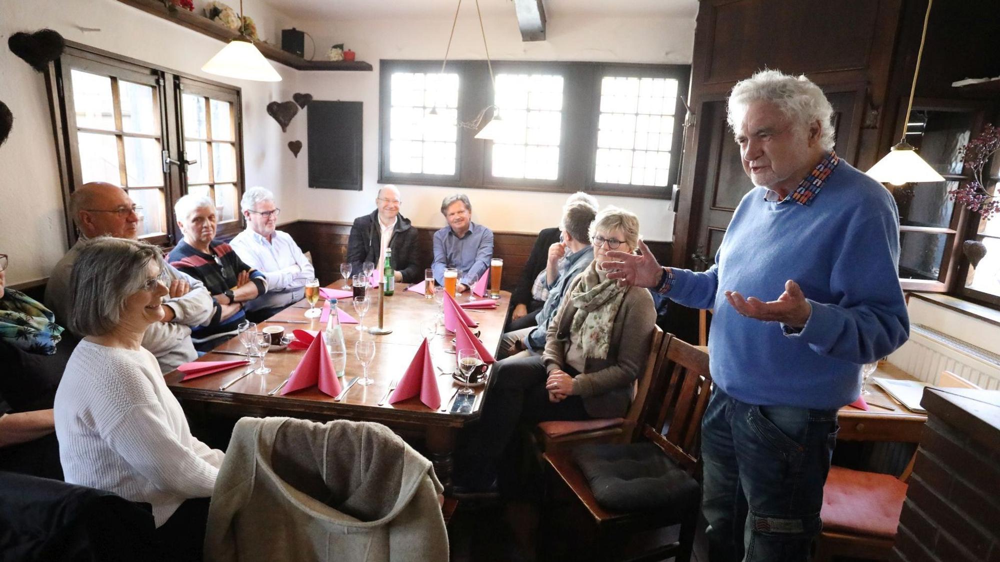 Ein Mann mit weißen Haaren und einem blauen Pullover spricht vor Zuhörern in einer Gaststätte.