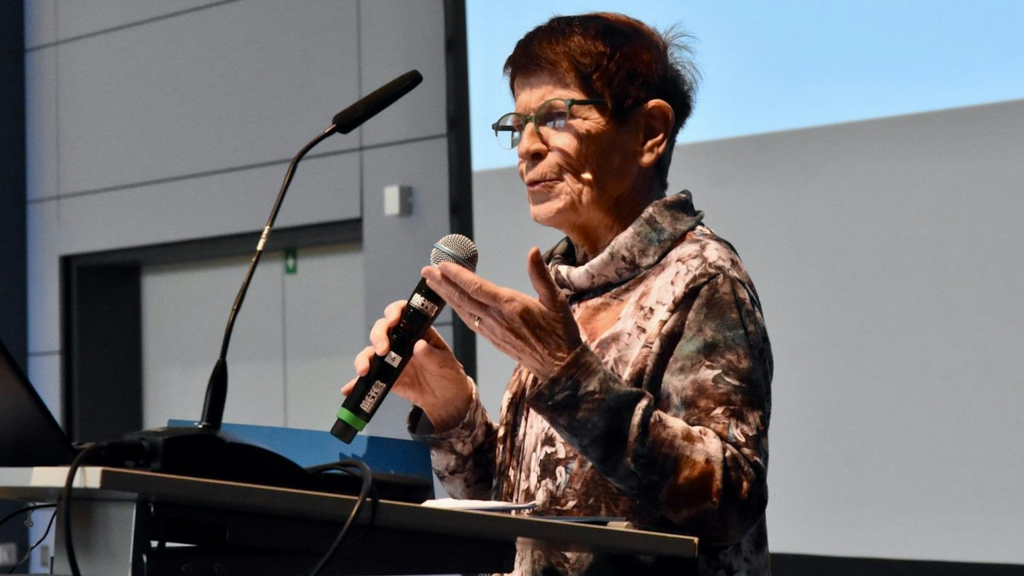 Eine Frau mit kurzen dunkelroten Haaren und einem Rollkragenpullover spricht mit Mikrofon an einem Rednerpult.