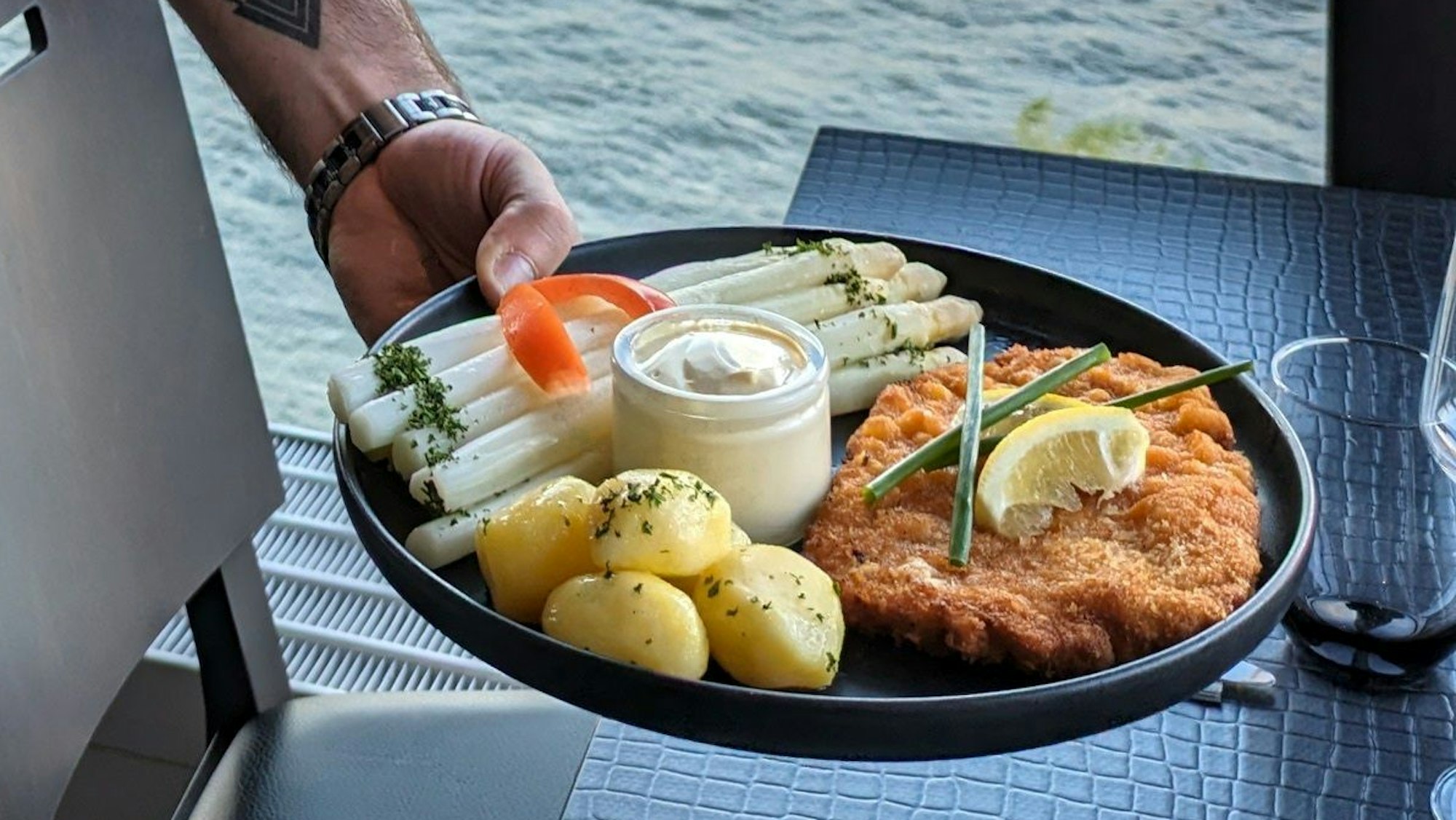 Ein Spargelgericht in der Hand eines Gastro-Mitarbeiters mit Blick durch ein Fenster auf den Rhein.