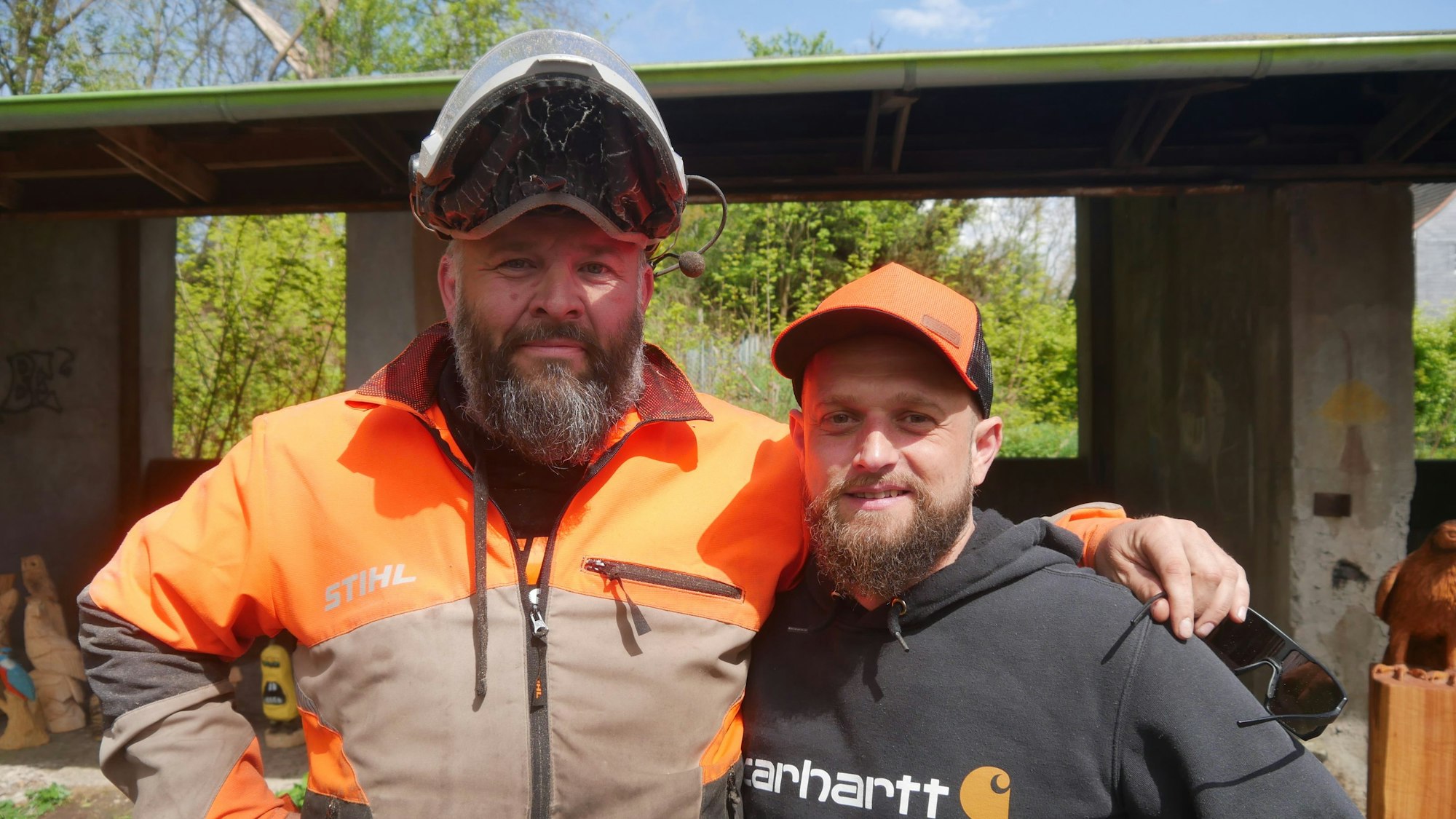 Zwei Männer; einer trägt einen schwarzen Kapuzenpulli und eine orange Schirmmütze, der andere einen Helm mit hochgeklapptem Visier.