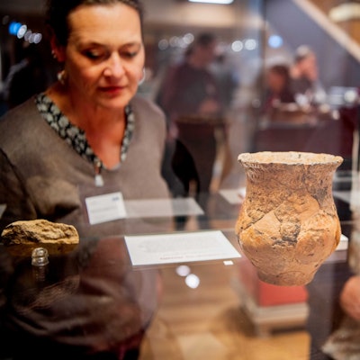 Das Bild zeigt einen Krug in einer Vitrine. Das Gefäß ist bei Großbüllesheim gefunden worden. Zwei Besucher schauen sich den Fund an.