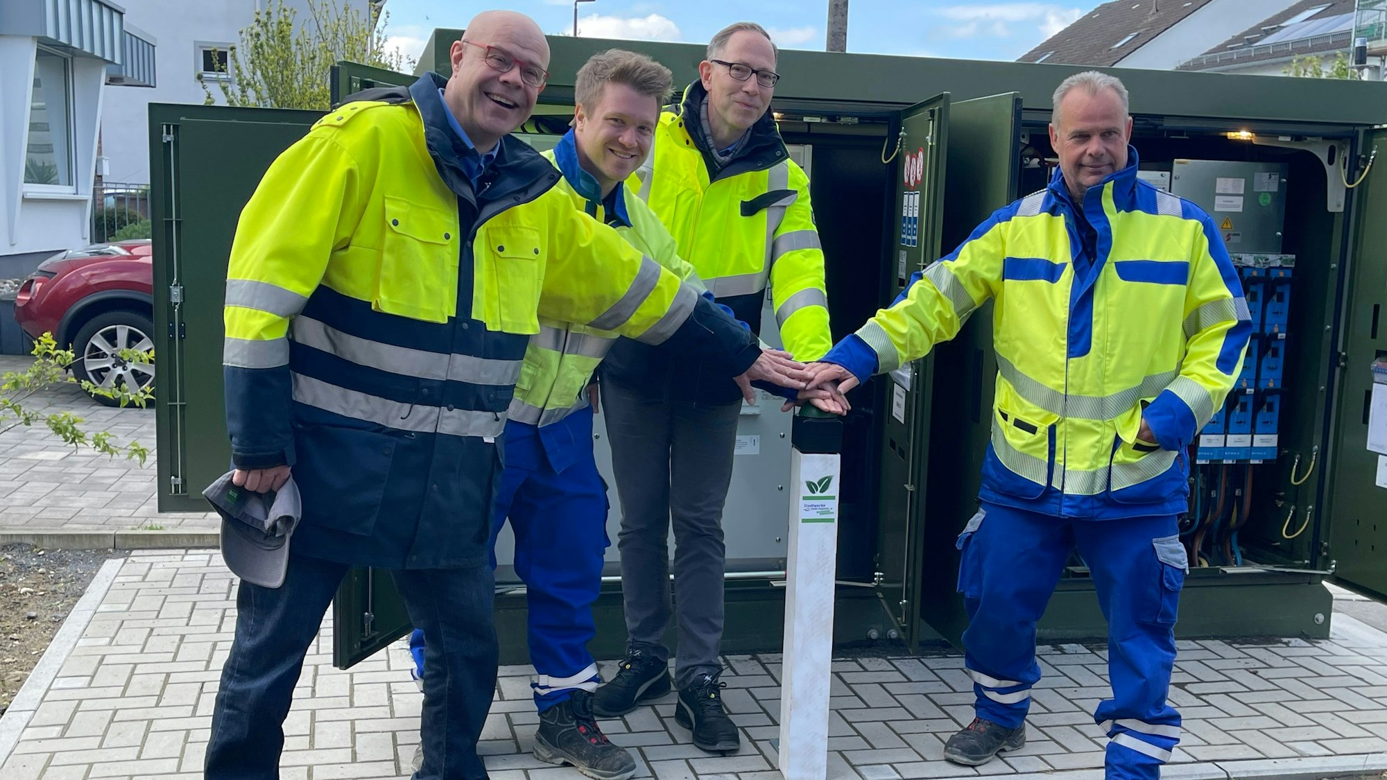 Vier Männer in blau-gelber Arbeitskleidung stehen vor einem grünen Kasten mit geöffneten Türen: der digitalen Strom-Ortsnetzstation in Sankt Augustin-Menden.
