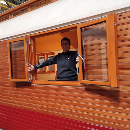 Eine Frau öffnet ein zweiflügeliges Fenster eines hölzernen Zirkuswagens.