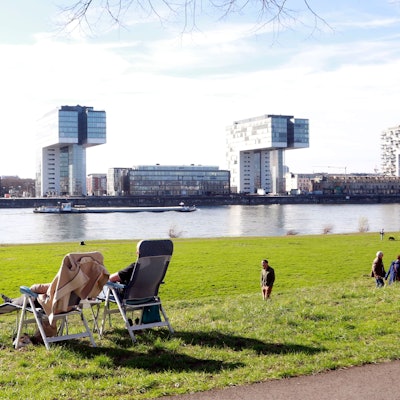 An der Alfred-Schütte-Allee, mit Panorama auf die Kölner Kranhäuser, liegen zwei auf ihren Sonnenliegen und genießen den Ausblick.