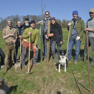 Tatkräftig packen Vorstand Patrick Beyer und die Ehrenamtler beim Pflanzen der Obstbäume auf dem Agro-Forst an. Die Gruppe steht auf einer Wiese beisammen. Alle haben einen Spaten in der Hand.