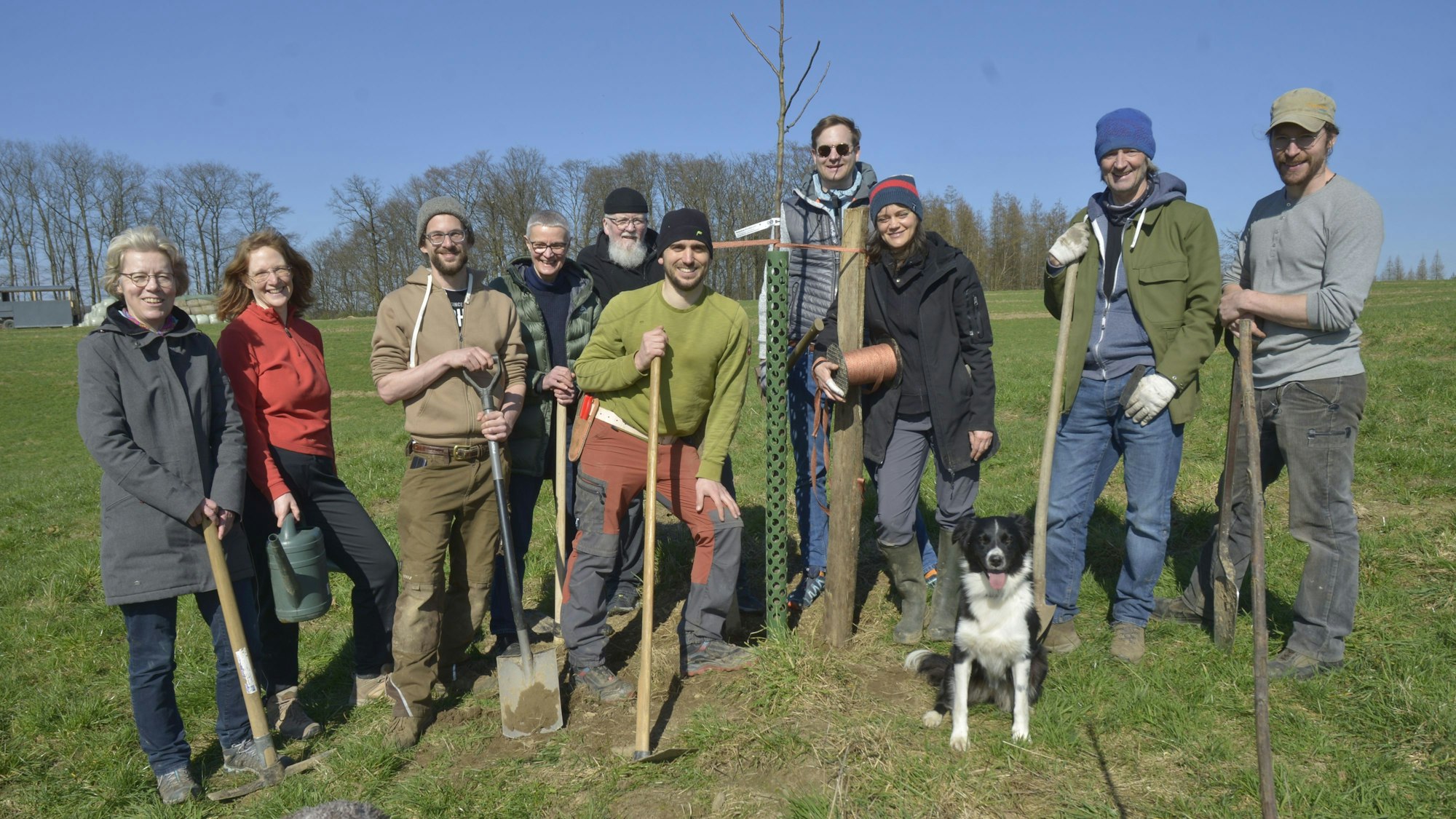 Tatkräftig packen Vorstand Patrick Beyer und die Ehrenamtler beim Pflanzen der Obstbäume auf dem Agro-Forst an. Die Gruppe steht auf einer Wiese beisammen. Alle haben einen Spaten in der Hand.