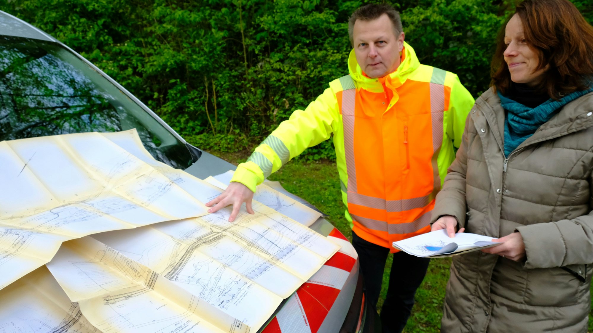 Mario Dittmann und Ines Rick stehen vor einem Auto, auf deren Motorhaube Pläne liegen. Dittmann zeigt darauf.