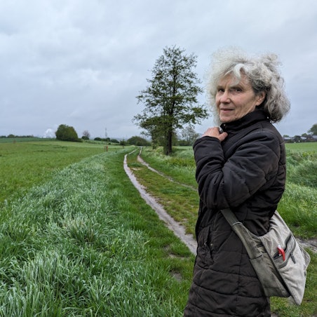 Die Ratsfrau Inge Cürten-Noack von den Grünen steht auf einem Feldweg vor einem Ackerstreifen, auf dem junge Bäume gepflanzt worden sind.