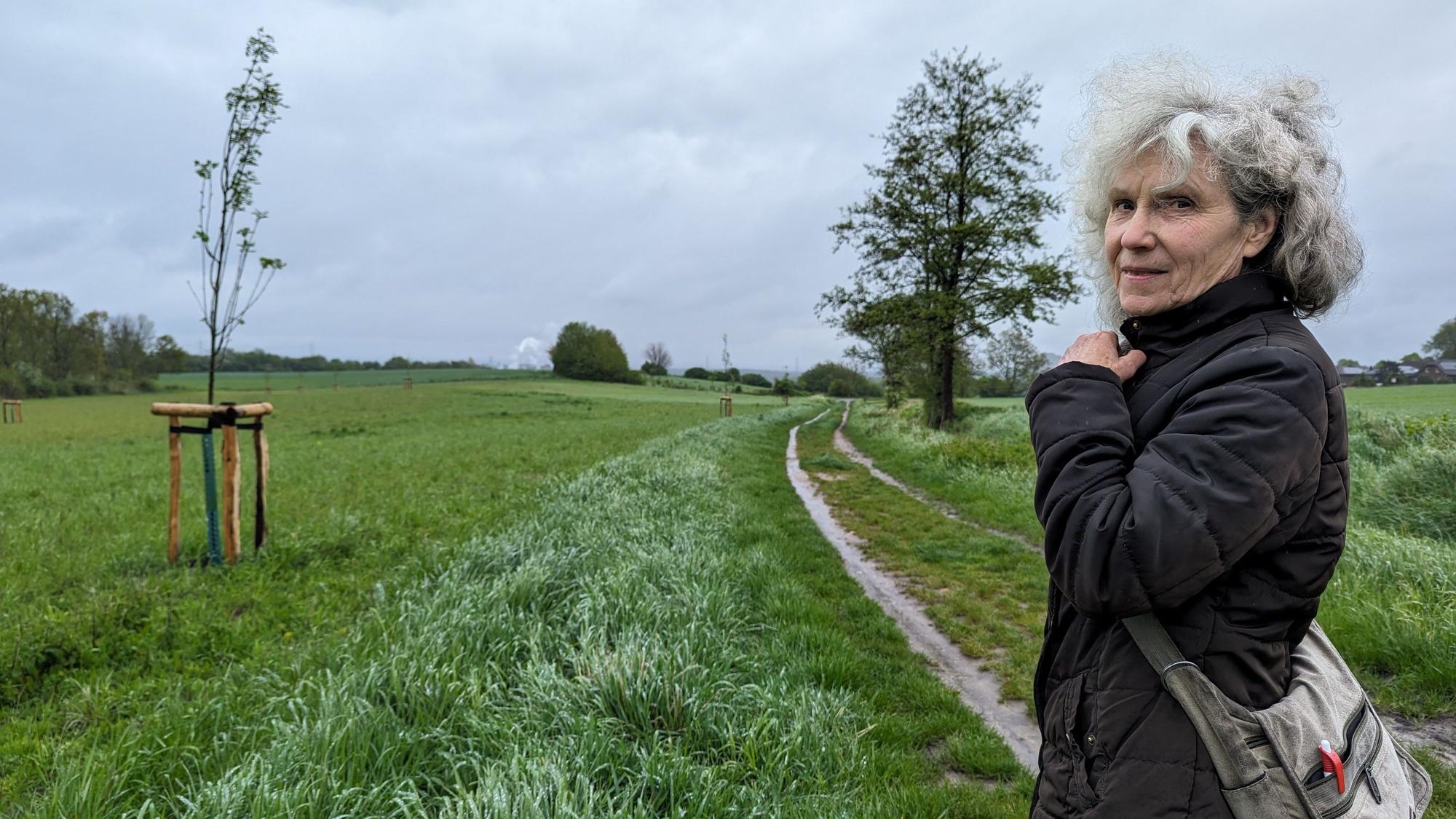Die Ratsfrau Inge Cürten-Noack von den Grünen steht auf einem Feldweg vor einem Ackerstreifen, auf dem junge Bäume gepflanzt worden sind.